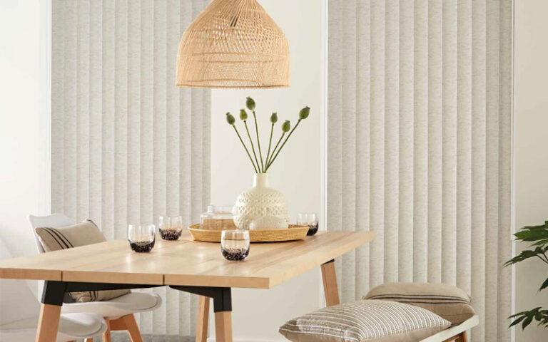 Textured cream vertical blinds behind a wooden dining table with a woven pendant light
