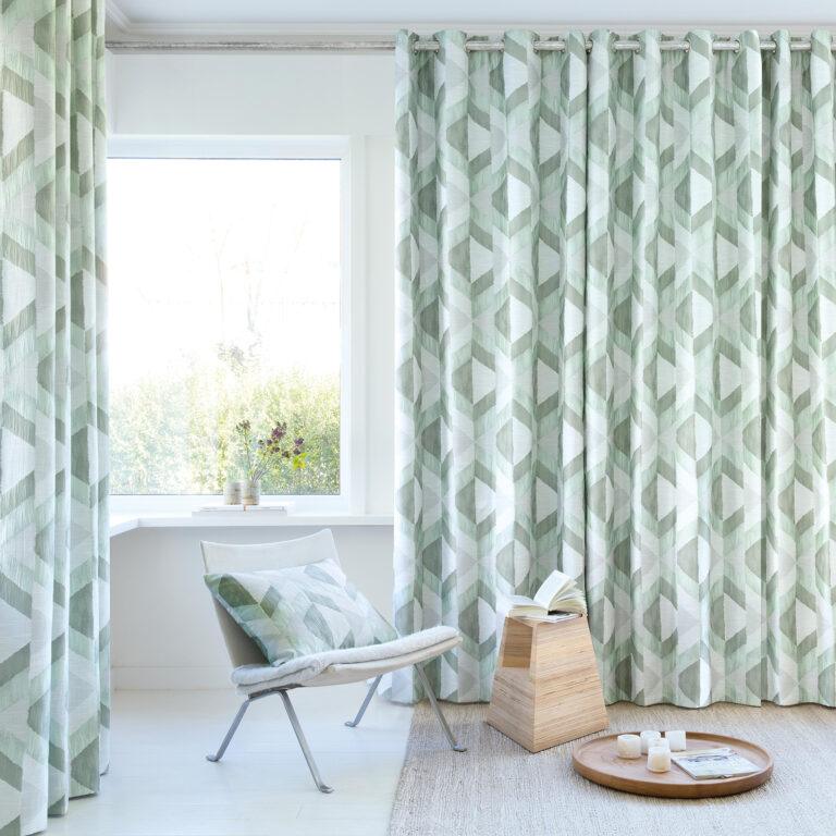 Green and white geometric eyelet curtains on a pole beside a window with a chair and side table